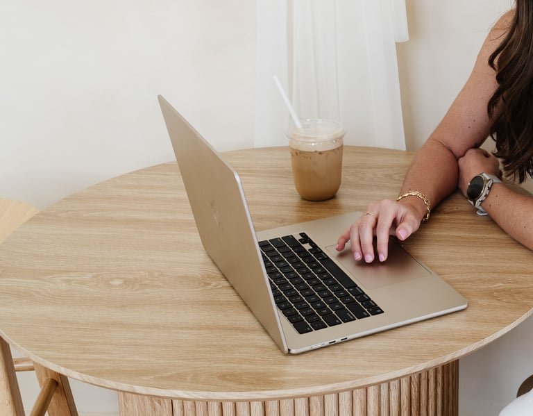 Person working on laptop at wooden desk with iced coffee drink in bright, minimalist workspace
